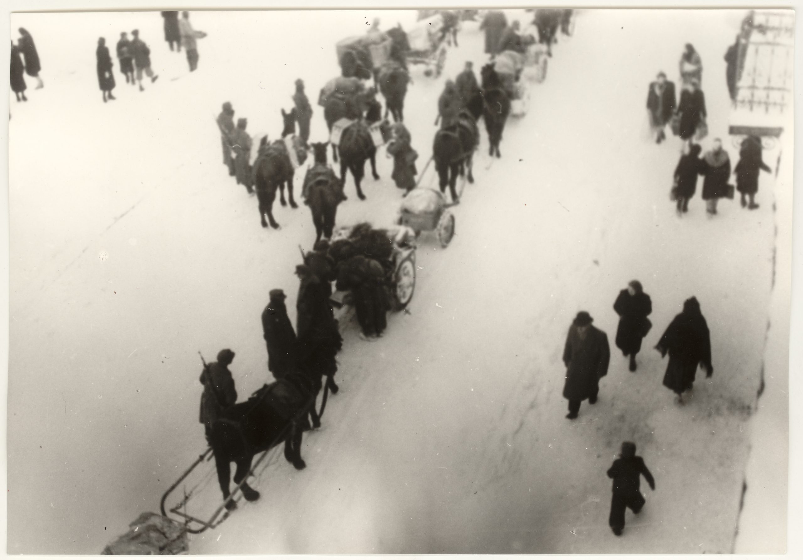 Fotografia czarnobiała przedstawia ludzi spacerujących podczas zimy. Zdjęcie zrobione jest z lotu ptaka