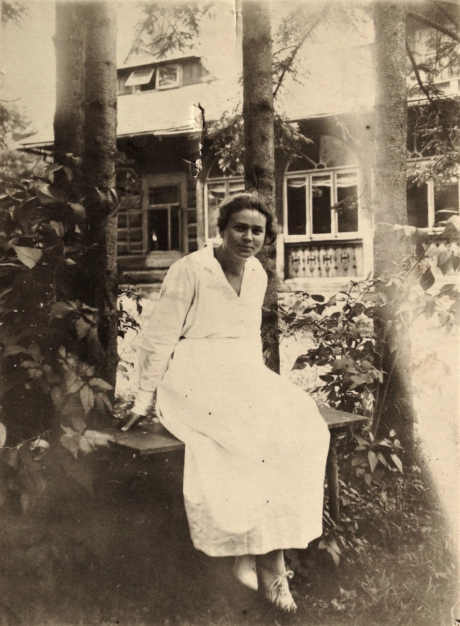 Fotografia czarno-biała. Przedstawia kobietę siedzącą na ławce. Z nią znajduje się drewniany dom